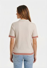 Mujer con cabello largo y castaño, vestida con un top beige de manga corta con ribetes a rayas rojas en los puños y el dobladillo, posando frente a un fondo blanco.