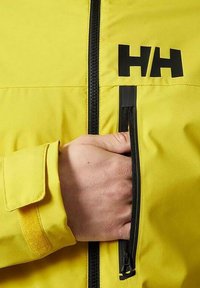 Giacca impermeabile gialla con zip nera, logo "HH," polsini in velcro e una tasca laterale per un sicuro stoccaggio. Tessuto resistente e cuciture curate.