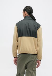 Fleece bunda s černým nylonovým horním dílem, béžovou spodní částí, elastickým pasem a dlouhými rukávy. Vyznačuje se volným střihem a jedinečným barevným blokováním.