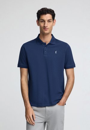 Polo shirt bleu marine en tissu lisse, avec un col à trois boutons et un petit emblème blanc sur la poitrine gauche. Manches courtes.