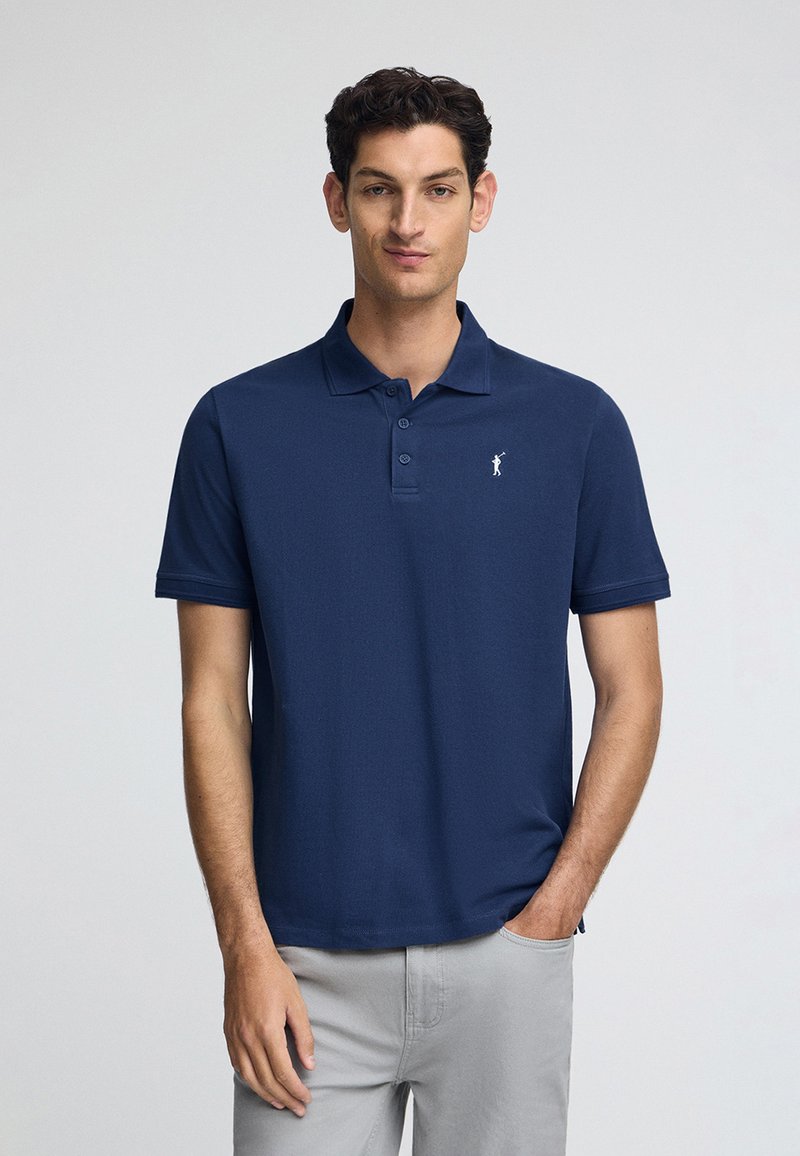 Mörkblå pikétröja av slät tyg med tre knappar på kragen och ett litet vitt emblem på vänster bröst. Kort ärm.