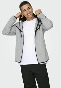 Chaqueta de cremallera gris claro con ribetes negros y dos bolsillos frontales con cremallera, llevada sobre una camisa blanca. Cuenta con capucha y textura suave.