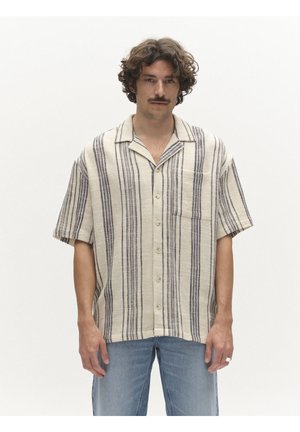 Kitsate juuste ja vuntsidega mees, kes kannab beeži lühikeste varrukatega nööbitavat särki vertikaalsete mustade triipudega ja siniseid teksapükse, seisab valgel taustal.