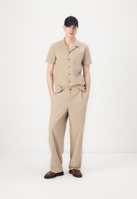 Camisa beige de manga corta con botonera y un bolsillo en el pecho, acompañada de pantalones sueltos en beige. Lleva una gorra negra y zapatos marrones.