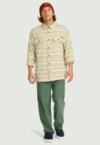 Homem em um gorro vermelho, camisa de botão bege com padrão, calças verdes e ténis escuros de pé contra um fundo simples.