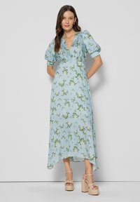 Jasnoniebieska sukienka w kwiaty z bufkami, dekoltem w serek i asymetrycznym dołem. Materiał ozdobiony wzorem zielonych liści. Noszona z beżowymi butami na platformie.