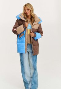 Žena oblečená v hnedej a modrej prešívanej bunde cez sveter s modrým designom huby a vo voľných, vyblednutých modrých džínsoch, stojí na bielom pozadí.
