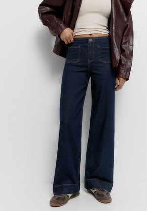 Personne portant un jean large taille haute bleu foncé, un débardeur beige, une veste en cuir bordeaux et des baskets marron, debout devant un fond blanc.