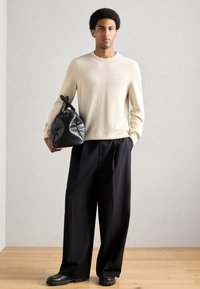 Suéter de punto beige, pantalones negros holgados, bolso de cuero negro con textura y zapatos negros gruesos. Fondo liso, suelo natural de madera.