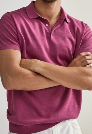 Mann mit verschränkten Armen, der ein kurzärmeliges pinkfarbenes Poloshirt und weiße Hosen vor einem schlichten hellen Hintergrund trägt.