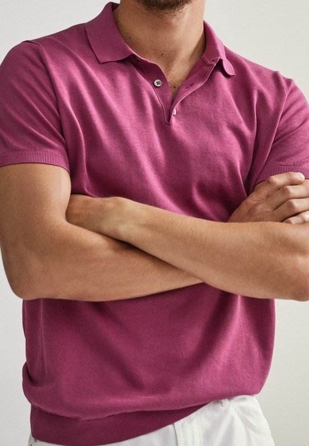Homme les bras croisés portant un polo rose à manches courtes et un pantalon blanc sur un fond clair uni.