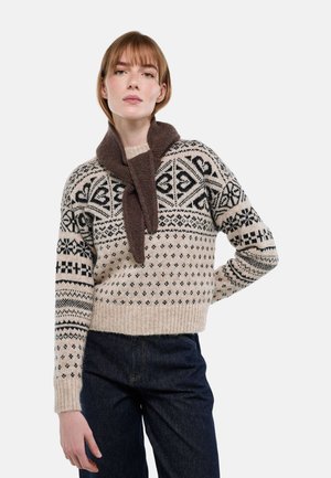 Femme portant un pull beige à motifs noirs, une écharpe sombre nouée autour du cou et un jean en denim foncé, debout avec une main sur la hanche.