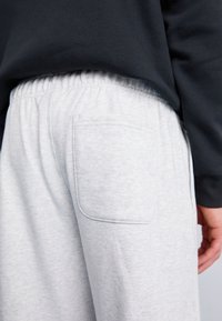 Šviesiai pilki sportiniai kelniukai su užpakaline kišene ir elastingu juosmeniu, pagaminti iš minkštos medžiagos su subtilia tekstūra. Artimas šoninis vaizdas.