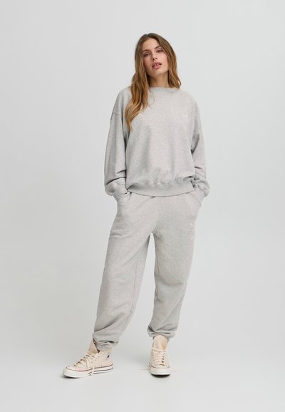 Felpa in cotone grigio chiaro con vestibilità loose fit e polsini a coste, abbinata a pantaloni jogger coordinati. Dettaglio del logo bianco sul lato sinistro del petto. Sneakers beige.