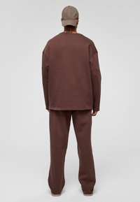 Bruine sweatshirt en bijpassende broek van zacht materiaal, met een relaxte pasvorm en minimale naden. Achteraanzicht met een baseballpet.