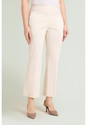 Femme portant un pantalon droit couleur crème et des escarpins slingback taupe à bout pointu, debout devant un fond uni.