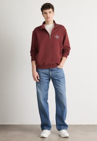 Μπορντό sweatshirt με φερμουάρ στο λαιμό και λεπτομέρεια λογότυπου, συνδυασμένο με ανοιχτό μπλε χαλαρά παντελόνια τζιν και λευκά αθλητικά παπούτσια. Λείο ύφασμα και χαλαρός σχεδιασμός.