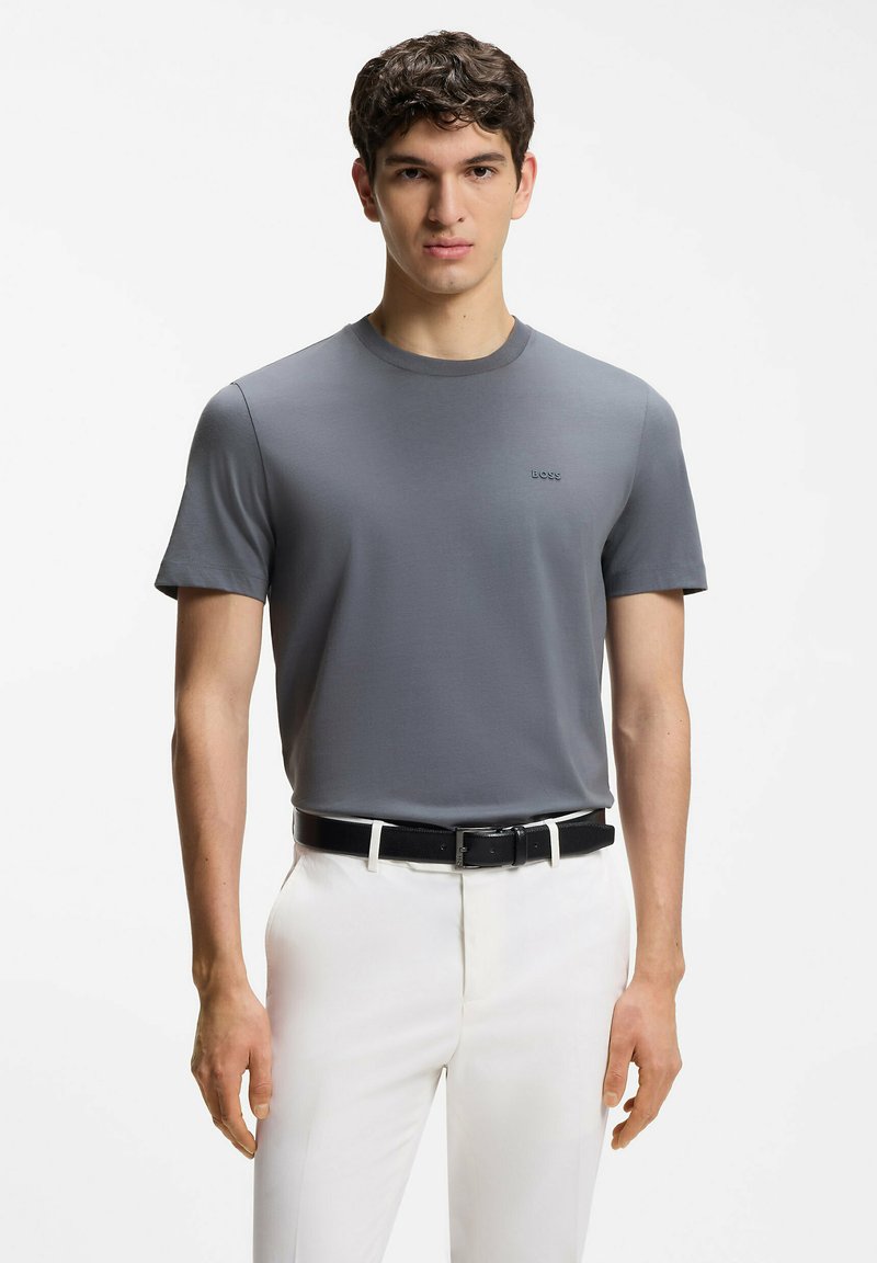 Jeune homme portant un t-shirt gris BOSS rentré dans un pantalon blanc avec une ceinture noire, se tenant devant un fond clair uni.