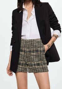 Blazer nero sopra una camicia bianca con bottoni, abbinato a pantaloni corti a quadri beige e verdi. Dettagli notevoli includono accenti con bottoni e tasche.