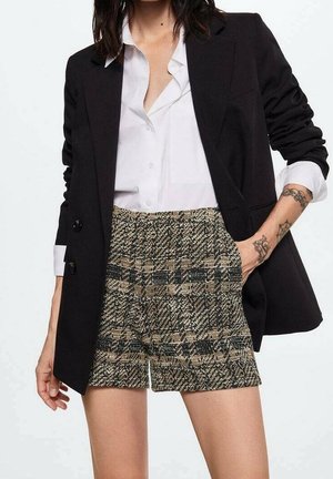 Blazer nero sopra una camicia bianca con bottoni, abbinato a pantaloni corti a quadri beige e verdi. Dettagli notevoli includono accenti con bottoni e tasche.