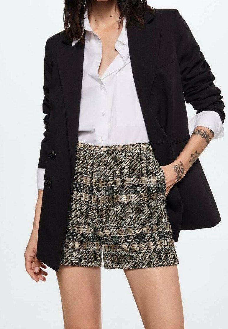 Blazer nero sopra una camicia bianca con bottoni, abbinato a pantaloni corti a quadri beige e verdi. Dettagli notevoli includono accenti con bottoni e tasche.