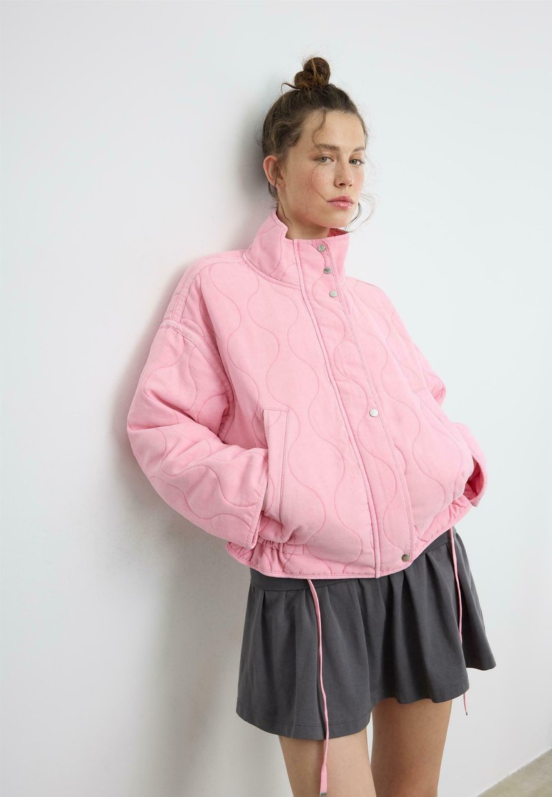 Jeune femme aux cheveux attachés en chignon portant une veste légère matelassée rose pâle et une jupe plissée foncée, appuyée contre un mur blanc.