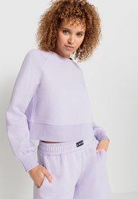 Felpa cropped lilla con collo rotondo, polsini e vita a coste. Abbinata a pantaloni corti in felpa coordinati con tasche laterali.