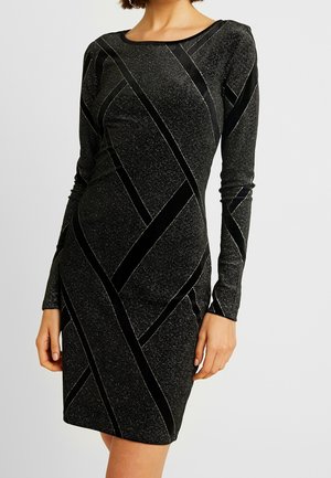 Schwarzes knielanges Kleid mit langen Ärmeln, diagonalen Samtstreifen und kleinen glitzernden Punkten, getragen von einer Person mit kurzen Haaren vor einfarbigem Hintergrund.