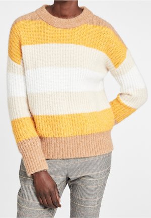 Persona que lleva un suéter de punto acanalado con rayas horizontales en amarillo mostaza, blanco, beige y camel, combinado con pantalones de cuadros.