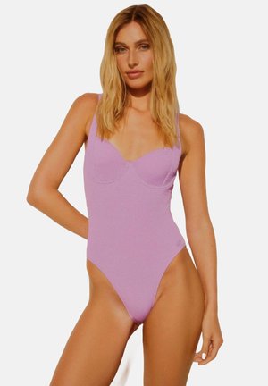 Jednoczęściowy kostium kąpielowy w kolorze lawendowym, z ryflowaną teksturą, głębokim dekoltem i szerokimi ramiączkami. Wysokie wycięcia nogawek podkreślają sylwetkę.