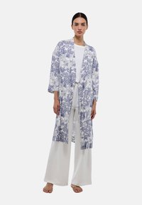 Kimono long à motif floral en tissu blanc et bleu, avec des manches larges, une ouverture sur le devant et une coupe décontractée, associé à un pantalon blanc.