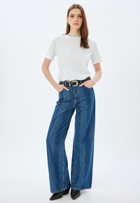 Camiseta blanca de manga corta combinada con unos jeans de mezclilla azul de pierna ancha con una textura sutil, llevada con un cinturón negro y zapatos negros de punta.