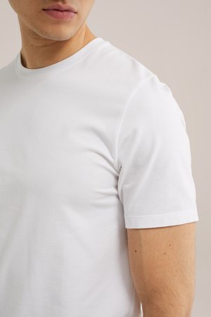 Gros plan d'une personne portant un t-shirt blanc uni à manches courtes avec un col rond sur un fond neutre.