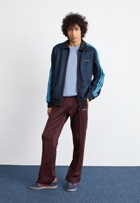 Marinblå träningsjacka med blå ränder, som matchas med vinröda träningsbyxor. Outfiten har en framficka och subtila varumärkesdetaljer.