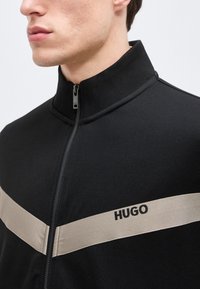 Κοντινό πλάνο ενός ατόμου που φοράει μαύρο μπουφάν με φερμουάρ, με μπεζ οριζόντια ρίγα και το κείμενο "HUGO" στο στήθος.