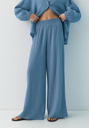 Person trägt lockere blaue strukturierte Hose, dazu passende offene Hemd und schwarze Sandalen, hebt das Hemd, um den nackten Bauch vor einem schlichten Hintergrund zu zeigen.