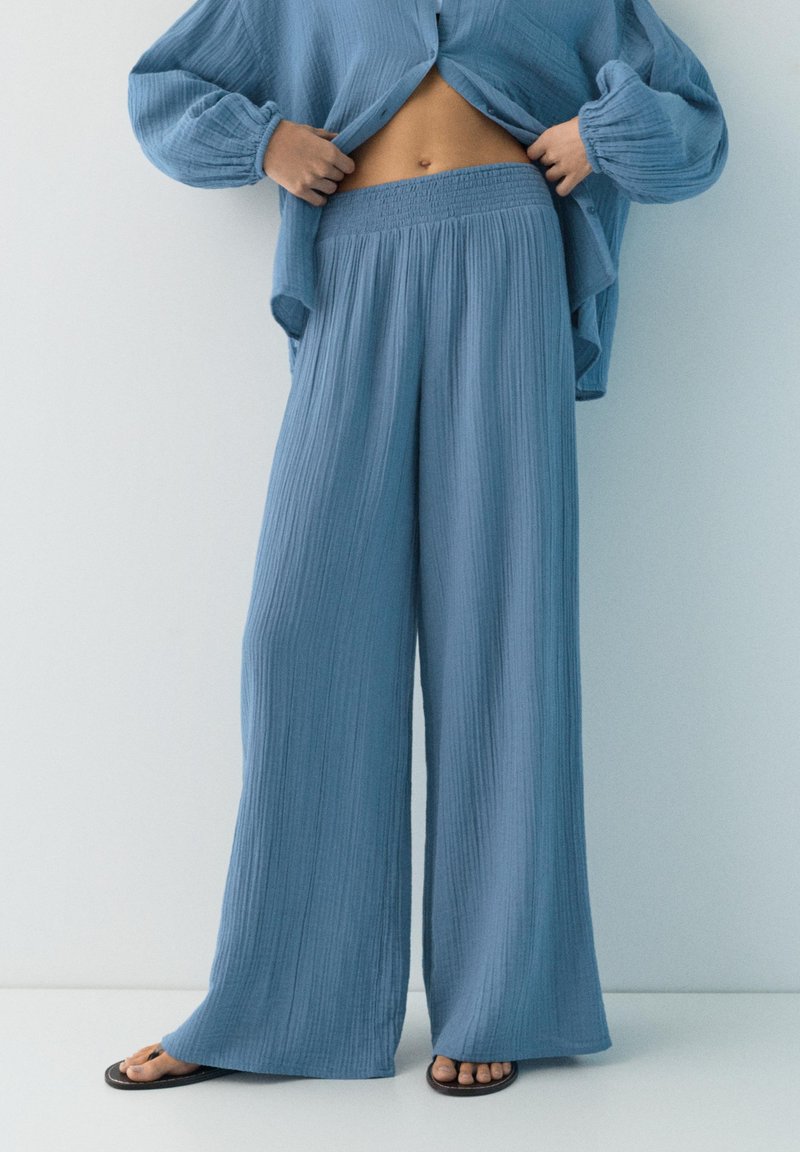 Osoba u širokim plavim hrapavim hlačama, usklađenoj otvorenoj košulji i crnim sandalama, podiže košulju kako bi otkrila prsa na jednobojnoj pozadini.