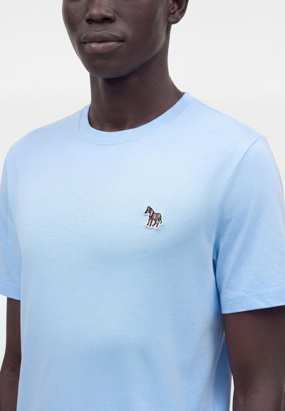 Άτομο που φορά ένα γαλάζιο t-shirt με στρογγυλή λαιμόκοψη και ένα μικρό, πολύχρωμο ριγωτό έμβλημα ζέβρας στο αριστερό στήθος.