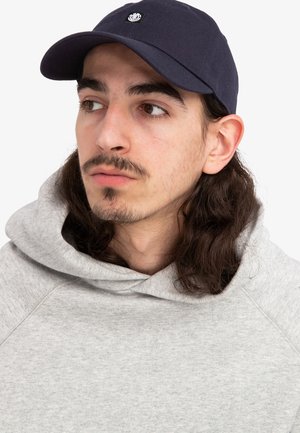 Casquette de marine avec un logo sur le devant, associée à un sweat à capuche gris clair. Le sweat a un tissu texturé et une coupe décontractée.