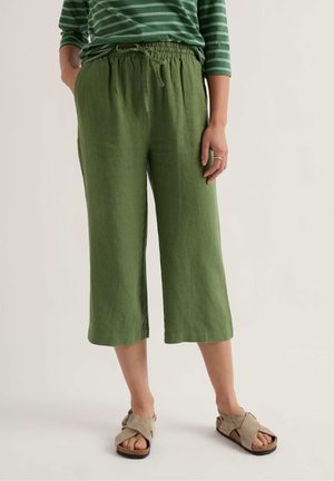 Persoană purtând pantaloni verzi scurți cu talie elastică și o cămașă cu mâneci lungi verde cu dungi, sandale bej pe un fundal simplu.