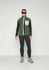 Colete de lã verde com detalhes em rosa, fecho de correr na frente e bolsos laterais, sobre mangas longas pretas e leggings. Usado com óculos de sol e ténis.