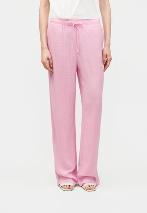 Άτομο που φοράει άνετο ροζ ανοιχτόχρωμο παντελόνι με κορδόνι στη μέση και λευκά σανδάλια ανοιχτά στα δάχτυλα, στέκεται μπροστά σε απλό φόντο.