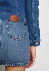 Niebieska kurtka dżinsowa z brązową skórzaną łatą, o dopasowanym kroju, z guzikami przy mankietach oraz detalami w postaci przeszyć na rękawach i u dołu.