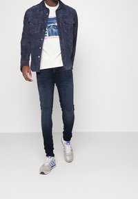 Chaqueta de camisa de ante azul marino, camiseta gráfica crema, jeans oscuros, zapatillas gris con acentos azules. Presenta dos bolsillos en el pecho y cierre de botones.