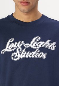 Tumedat sinine puuvillane T-särk, millel on valge tikandiga tekst "Low Lights Studios" kätksetud cursive-kirjas rinnal. Klassikaline ümara kaelusega disain.