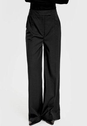 Czarne, szerokie spodnie z wysokim stanem i plisami z przodu, noszone z czarnymi, węższymi butami z ćwiekami na jasnym tle.