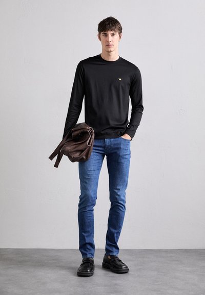 Chemise noire à manches longues avec un petit logo doré, associée à un jean bleu et des chaussures en cuir noires. Tient un sac en cuir marron.