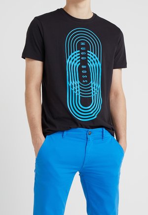 Osoba ubrana w czarny t-shirt Hugo Boss z niebieskimi koncentrycznymi owalnymi liniami i niebieskie spodnie, z rękami w kieszeniach, na tle jednolitego jasnego tła.