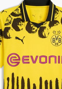 Tricou de fotbal galben cu modele grafice negre, guler în V negru, logo Puma pe pieptul stâng și inscriptionat "EVONIK" în roz.