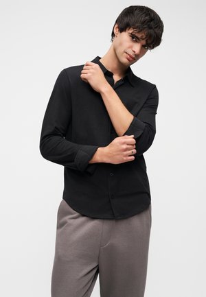 Sort skjorte med lange ærmer, knapper foran og opkrogede manchetter. Blødt stof, tætsiddende design. Kombineret med lysegrå bukser.